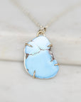 A close-up of a light blue turquoise stone pendant with natural matrix cracks, set in a gold-toned bezel and hanging from a silver-toned chain on a light marble surface.