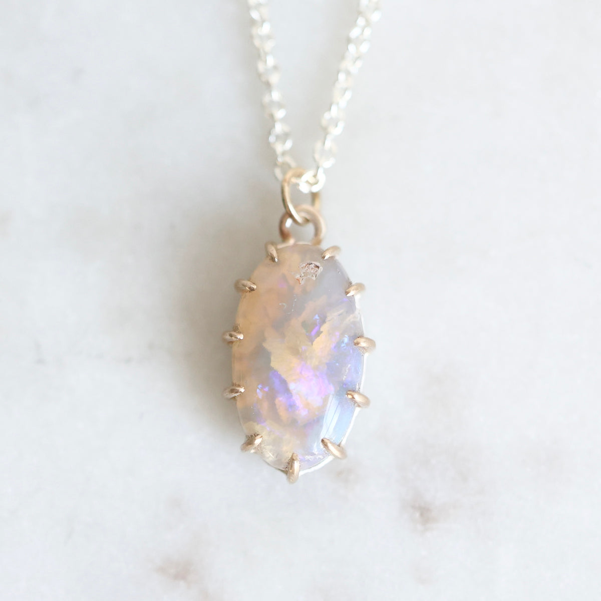 Purple and pink opal pendant set in yellow gold prongs against a marble background. 
