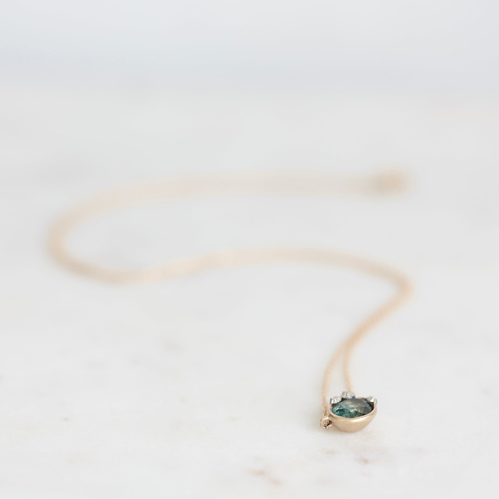A delicate gold necklace with a small oval pendant featuring a dark green stone, resting on a light marble surface.