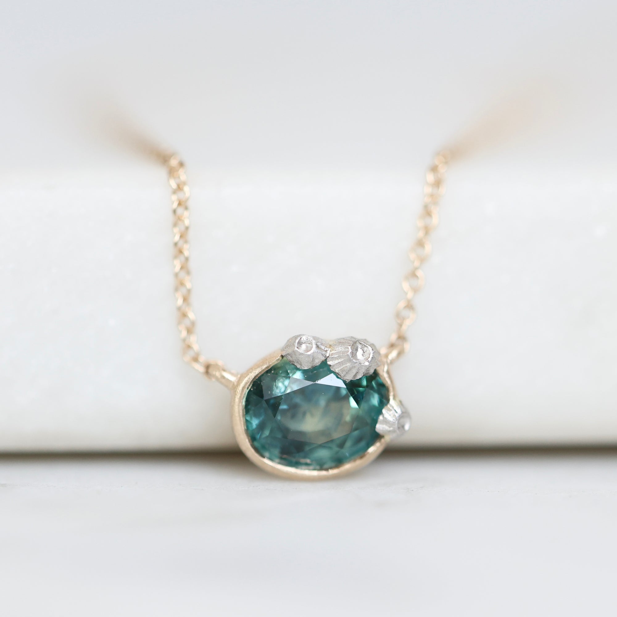 A delicate gold chain necklace with a pendant featuring a faceted teal-blue gemstone and small barnacles, displayed on a soft white background.