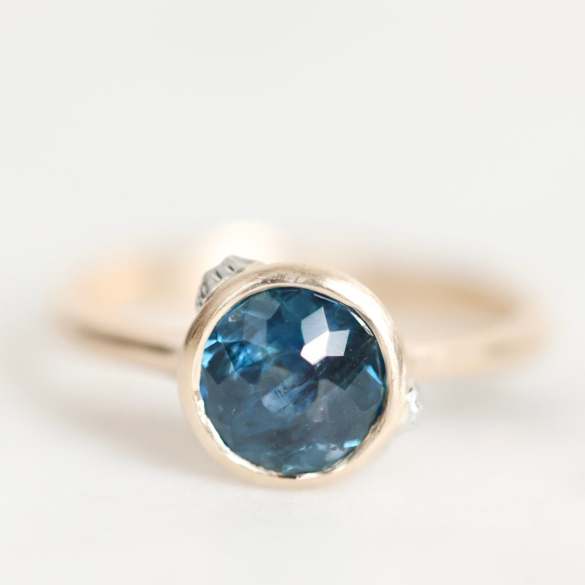 A close-up shot of a ring featuring a round-cut blue gemstone in a gold bezel setting, with a small clear accent stone on the side, set against a soft white background.