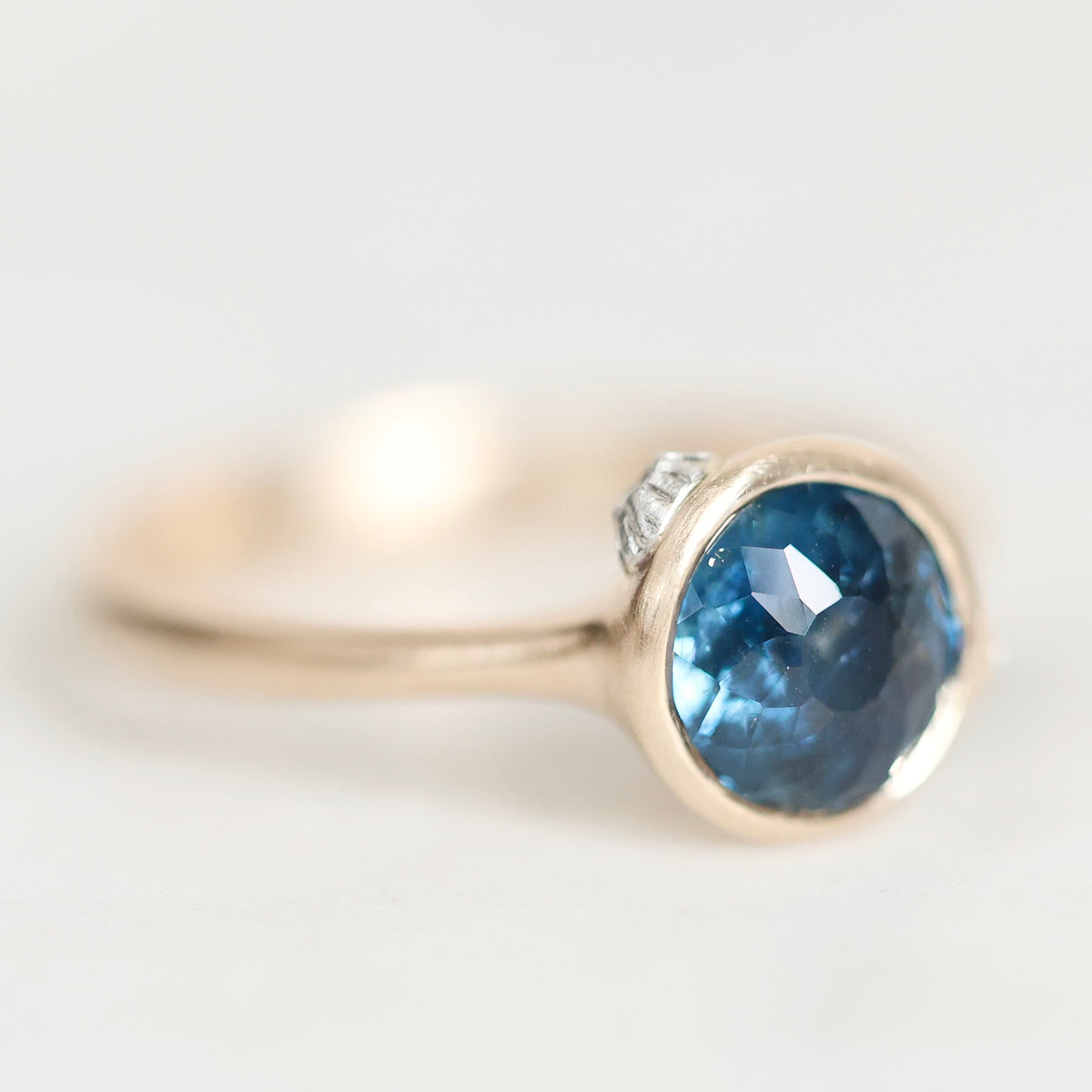 A close-up shot of a gold ring featuring a round-cut blue gemstone in a bezel setting, with a small clear accent stone on the side, set against a soft white background.