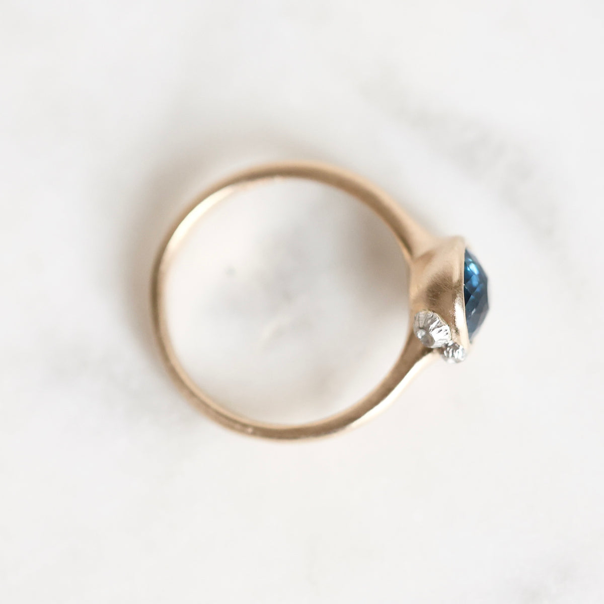 A gold ring with a small clear accent stone and a blue gemstone, photographed from above on a light marble surface.