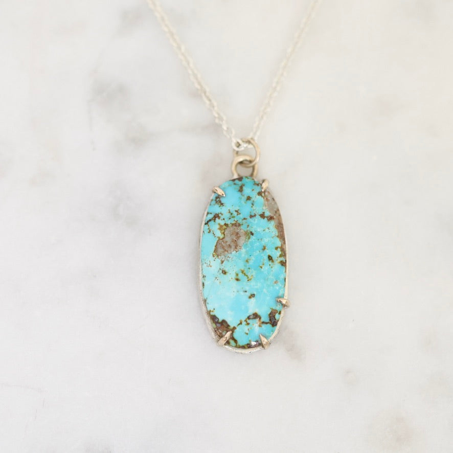 An oval turquoise pendant necklace with a silver bezel setting, resting on a white marble surface.