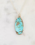 An oval turquoise pendant necklace with a silver bezel setting, resting on a white marble surface.