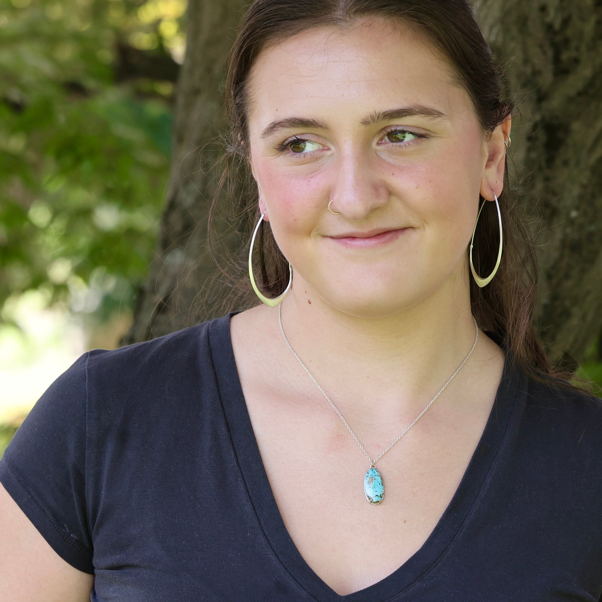 A woman outdoors wearing gold hoop earrings, a silver necklace with a blue-green stone pendant, and a dark navy blue v-neck t-shirt, with a tree trunk in the background.