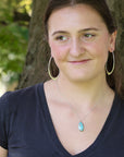 A woman outdoors wearing gold hoop earrings, a silver necklace with a blue-green stone pendant, and a dark navy blue v-neck t-shirt, with a tree trunk in the background.
