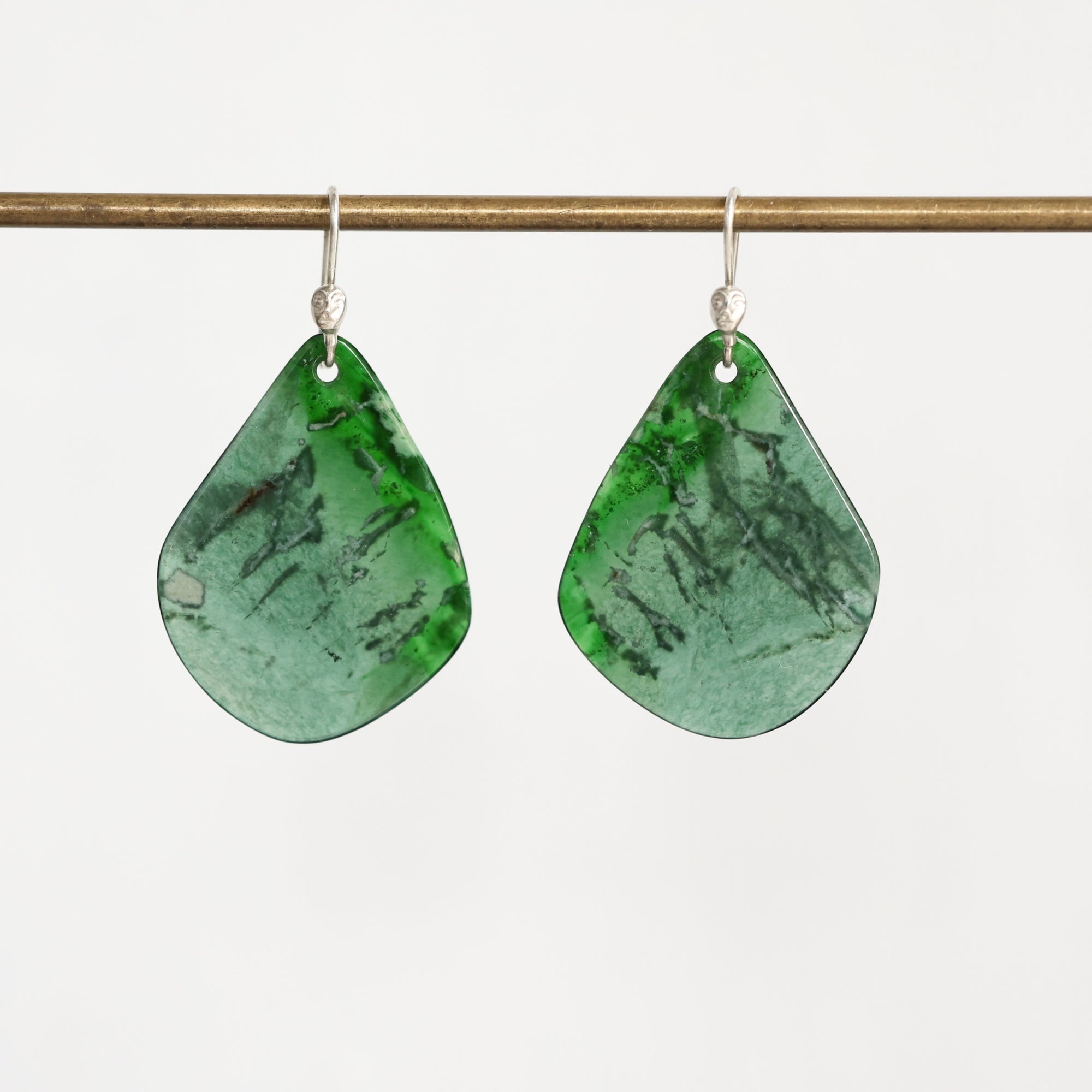 A pair of teardrop-shaped green stone dangle earrings with silver hooks, hanging from a thin horizontal brass rod against a plain light background.