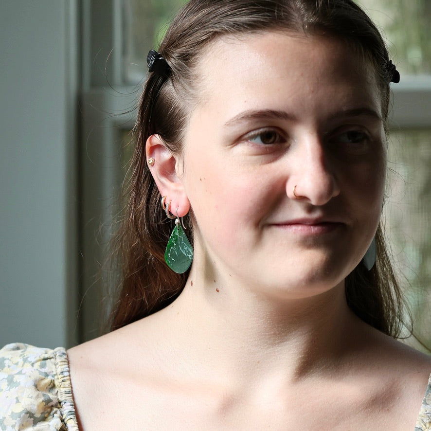 A close-up portrait of a woman wearing a pair of green teardrop-shaped dangle earrings with gold-toned studs, set against a soft-focus indoor background.
