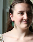 A close-up portrait of a woman wearing a pair of green teardrop-shaped dangle earrings with gold-toned studs, set against a soft-focus indoor background.