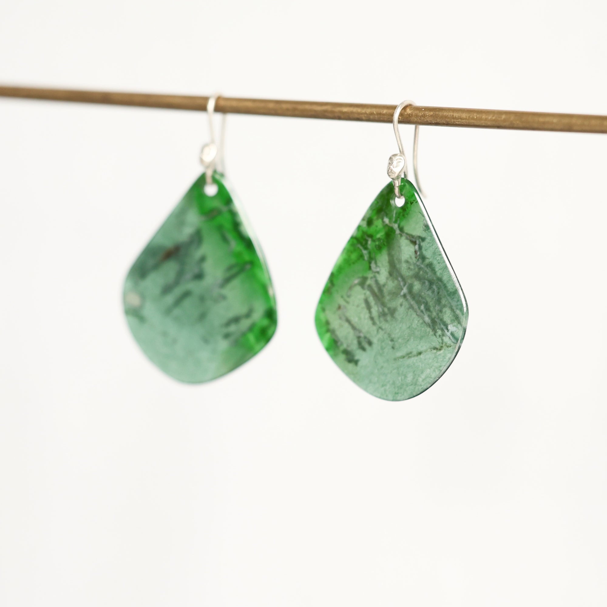 A pair of green teardrop-shaped dangle earrings with a marbled pattern, hanging from silver fishhook wires on a thin wooden rod against a plain white background.