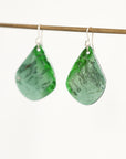 A pair of green teardrop-shaped dangle earrings with a marbled pattern, hanging from silver fishhook wires on a thin wooden rod against a plain white background.