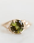 Green gemstone ring held in branchy gold prongs against a white background. 