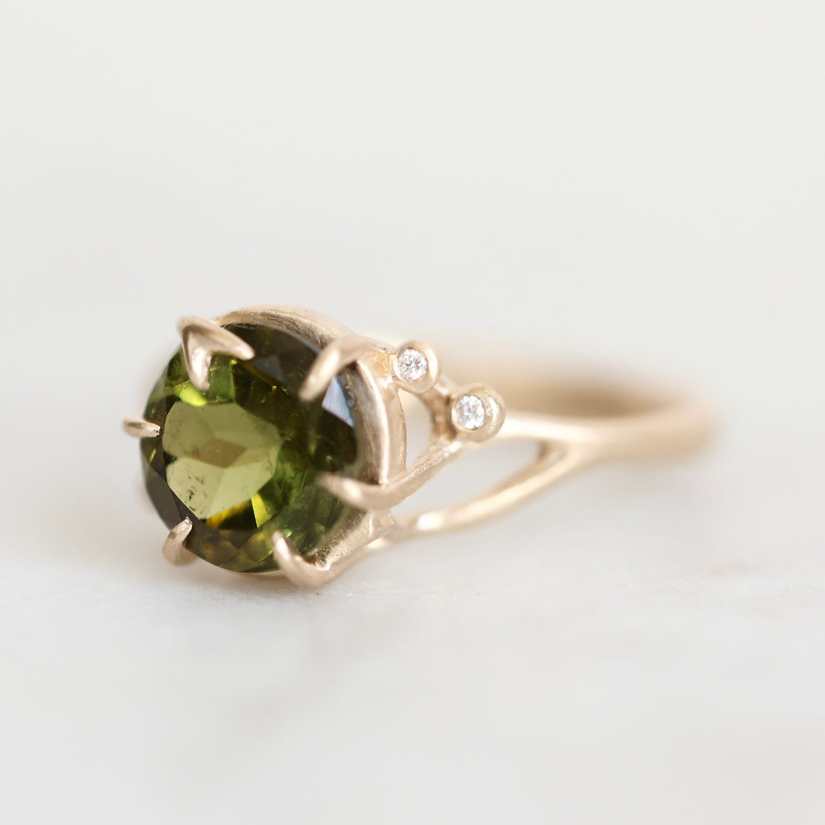 A yellow gold ring featuring a large oval green gemstone in a prong setting, accented with two small round-cut diamonds on the upper right side of the band, set against a clean white background.