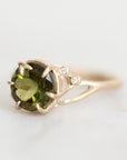 A yellow gold ring featuring a large oval green gemstone in a prong setting, accented with two small round-cut diamonds on the upper right side of the band, set against a clean white background.