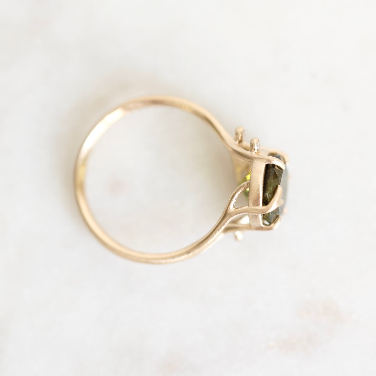 A gold ring with a bezel-set green gemstone, photographed from above on a light, off-white marble surface.
