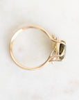 A gold ring with a bezel-set green gemstone, photographed from above on a light, off-white marble surface.
