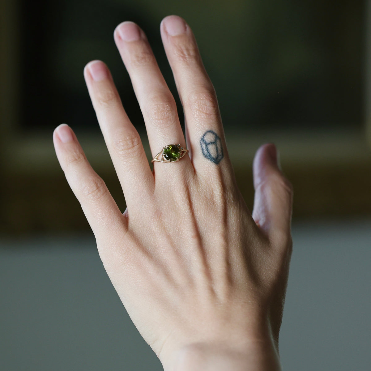 A close-up of a person's hand wearing a delicate gold ring with a green gemstone, set against a softly blurred dark background.