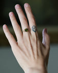 A close-up of a person's hand wearing a delicate gold ring with a green gemstone, set against a softly blurred dark background.
