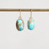 A pair of gold drop earrings with oval turquoise-blue stone pendants featuring natural brown and gold matrix patterns, hanging from a gold rod against a light gray background.