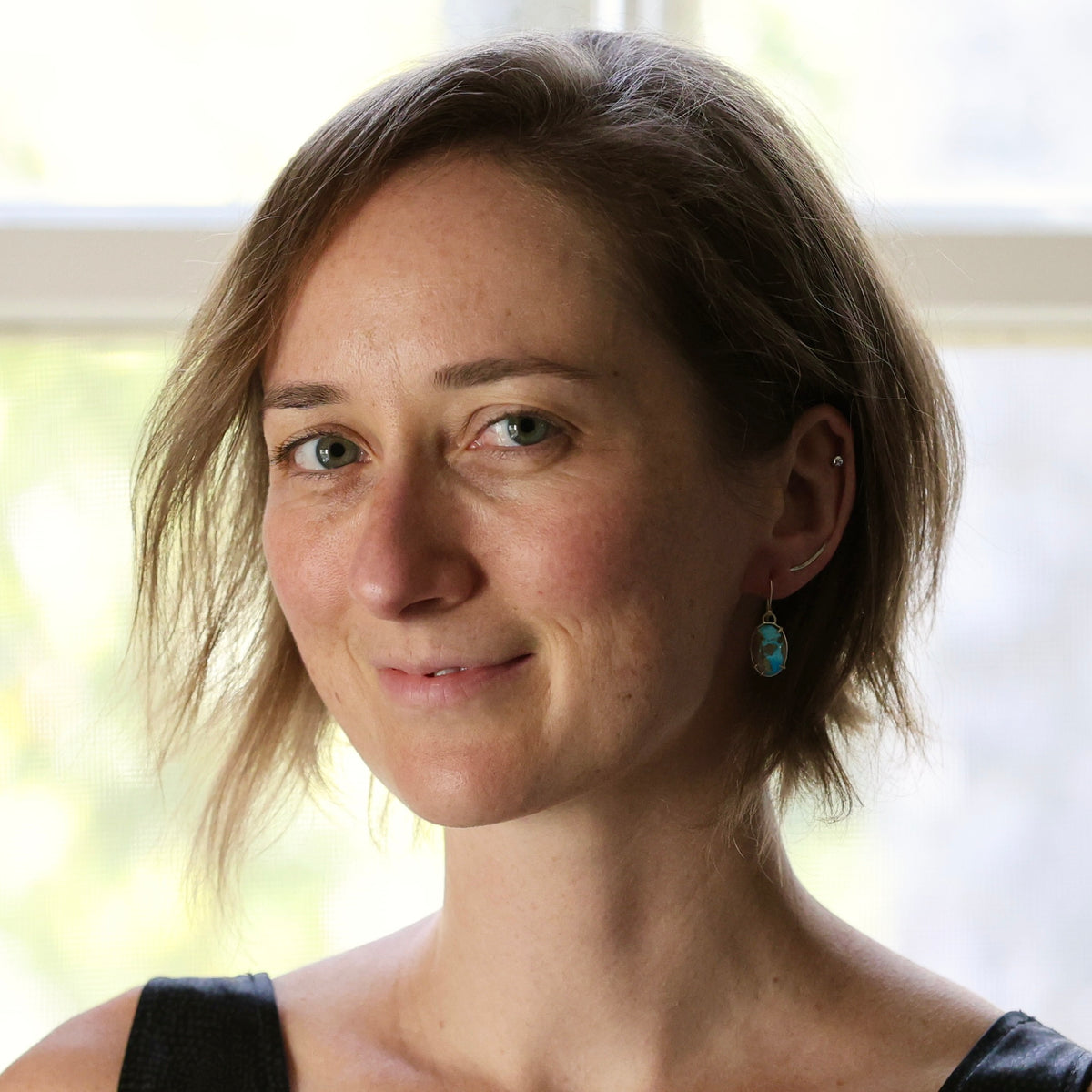 A close-up portrait of a woman with short brown hair, wearing a small turquoise earring, photographed in front of a softly blurred window background.