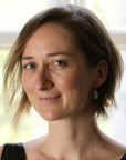 A close-up portrait of a woman with short brown hair, wearing a small turquoise earring, photographed in front of a softly blurred window background.