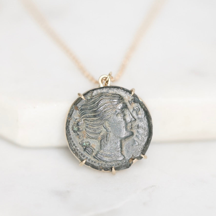 An ancient coin pendant necklace featuring a profile portrait of a woman, set in a gold-toned bezel and suspended from a delicate gold chain against a white marble background.