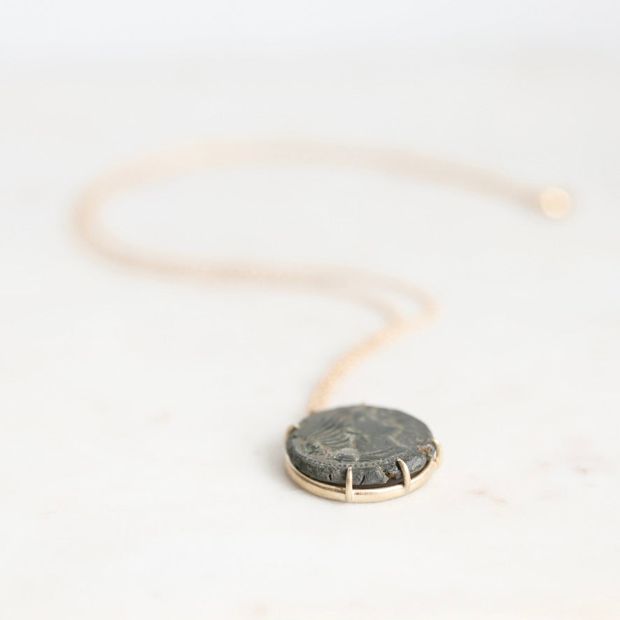 A delicate gold chain necklace with a round, dark gray stone pendant set in a gold bezel, resting on a light off-white marble surface.