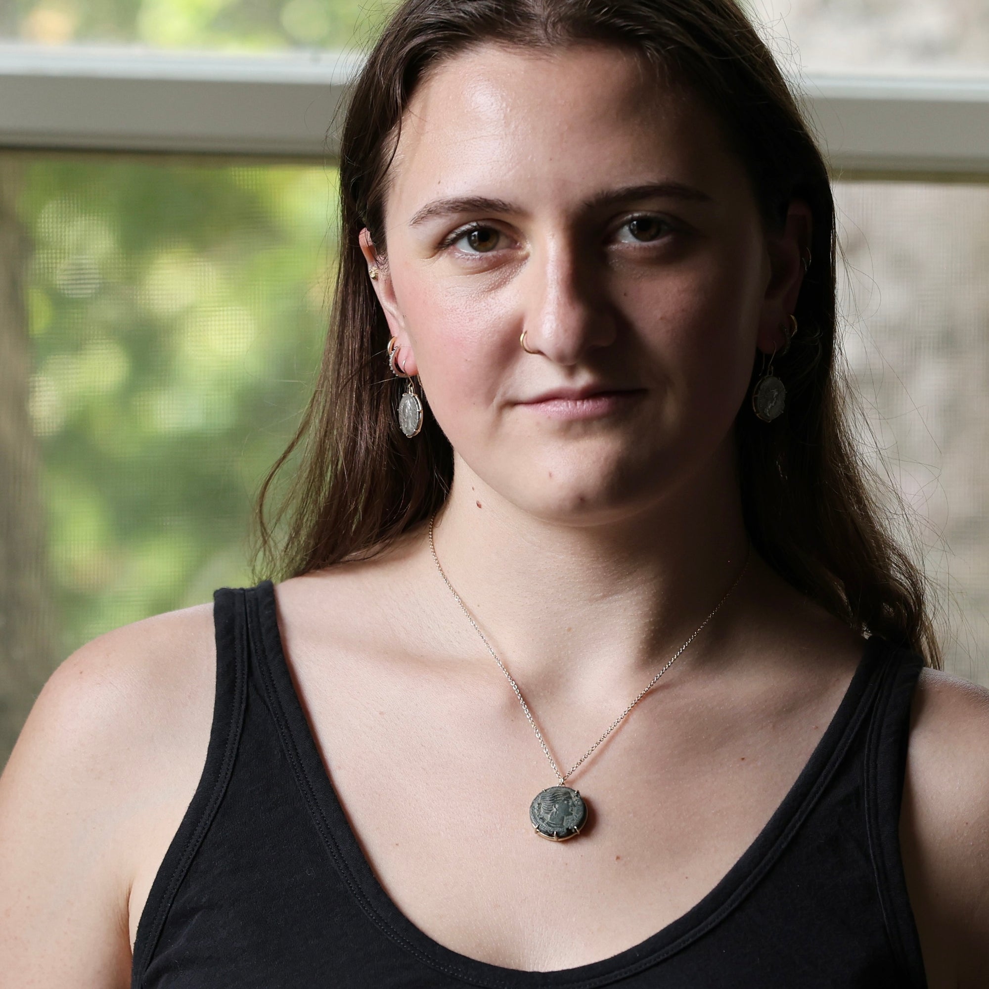 A woman with long brown hair wearing a black tank top, a silver pendant necklace with a round stone, and matching earrings, photographed in front of a softly blurred window.