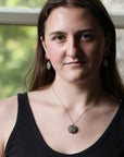 A woman with long brown hair wearing a black tank top, a silver pendant necklace with a round stone, and matching earrings, photographed in front of a softly blurred window.