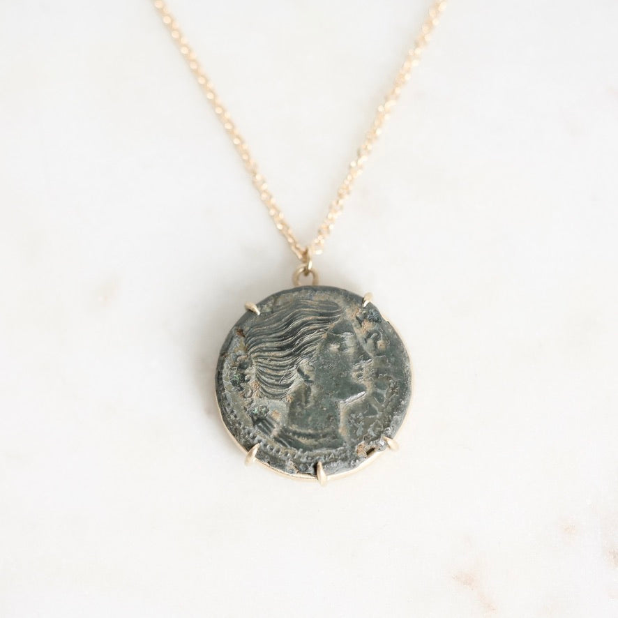 A gold chain necklace featuring a circular pendant made from an ancient, oxidized coin with a profile portrait, displayed on a light marble surface.