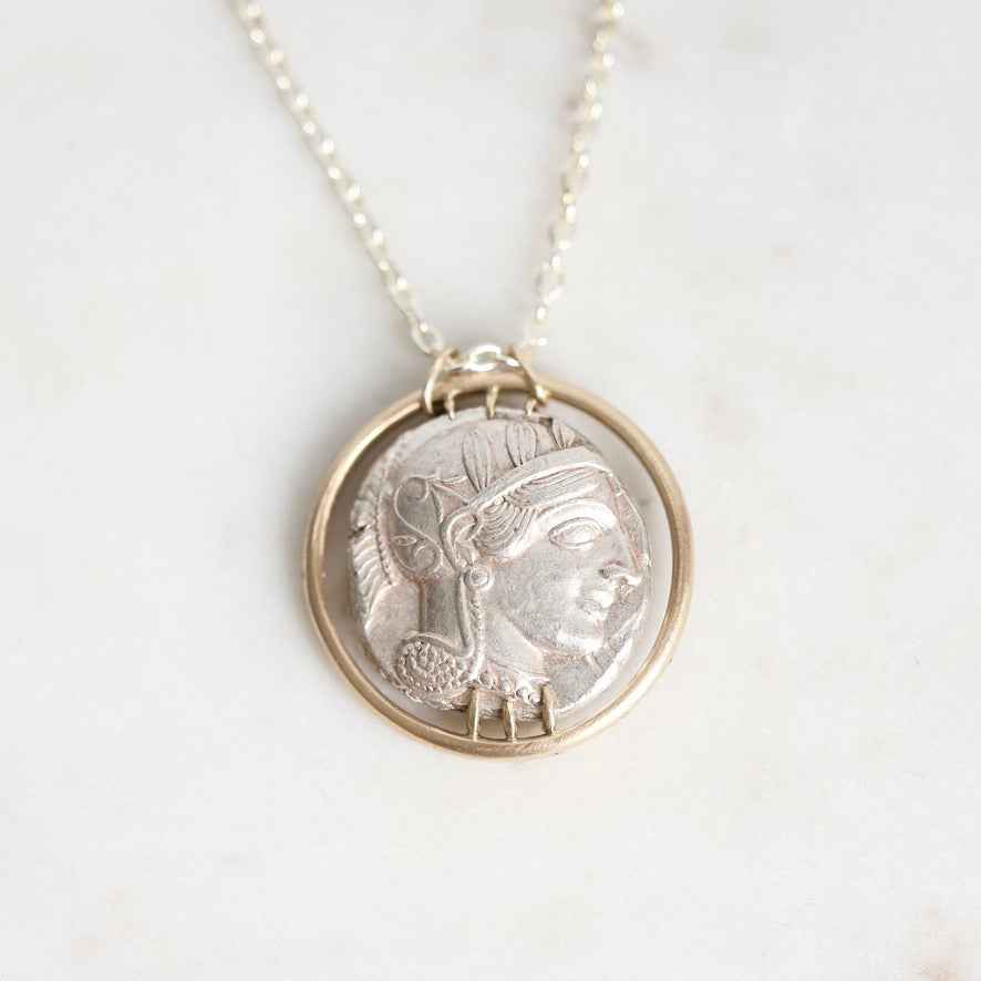A silver pendant necklace featuring an ancient Greek or Roman-style profile medallion, set in a gold-toned circular frame on a delicate silver chain, displayed on a light marble surface.