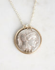 A silver pendant necklace featuring an ancient Greek or Roman-style profile medallion, set in a gold-toned circular frame on a delicate silver chain, displayed on a light marble surface.