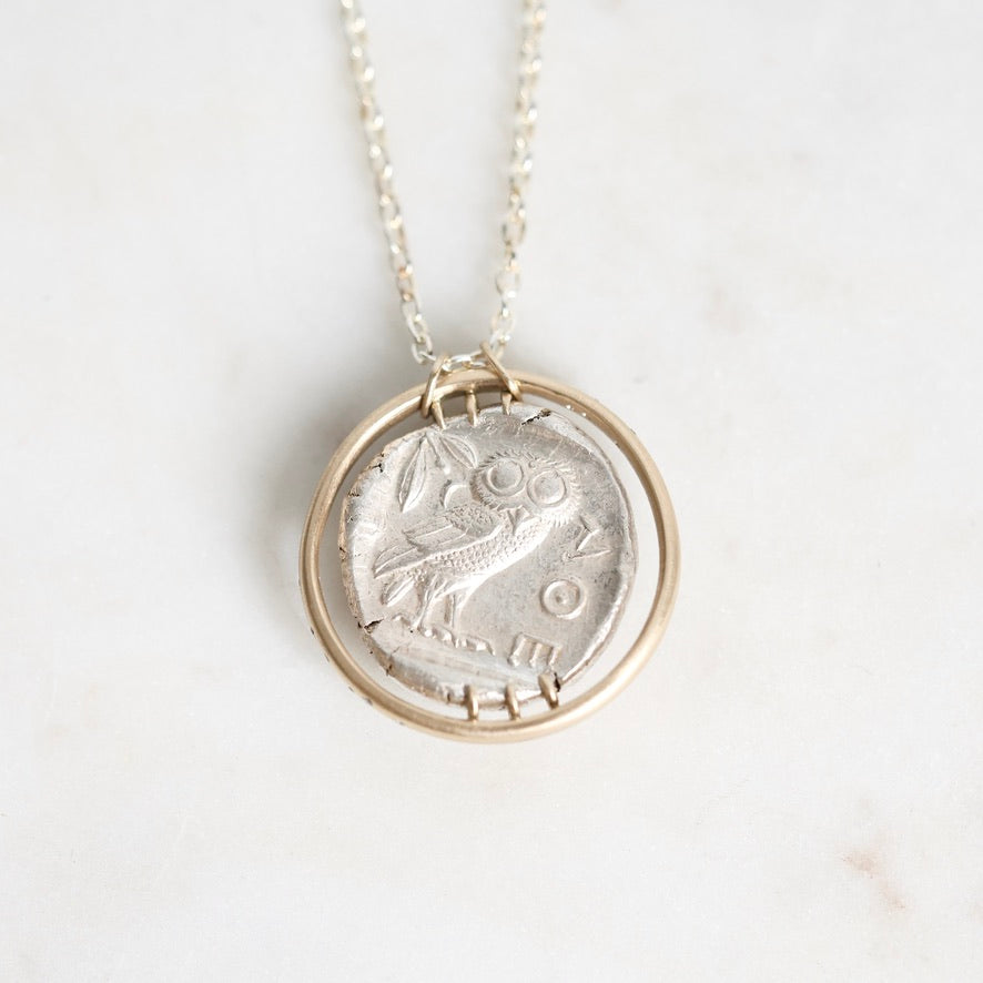 A silver pendant necklace featuring a circular coin-style charm with an engraved bird design, suspended from a delicate silver chain against a light marble background.