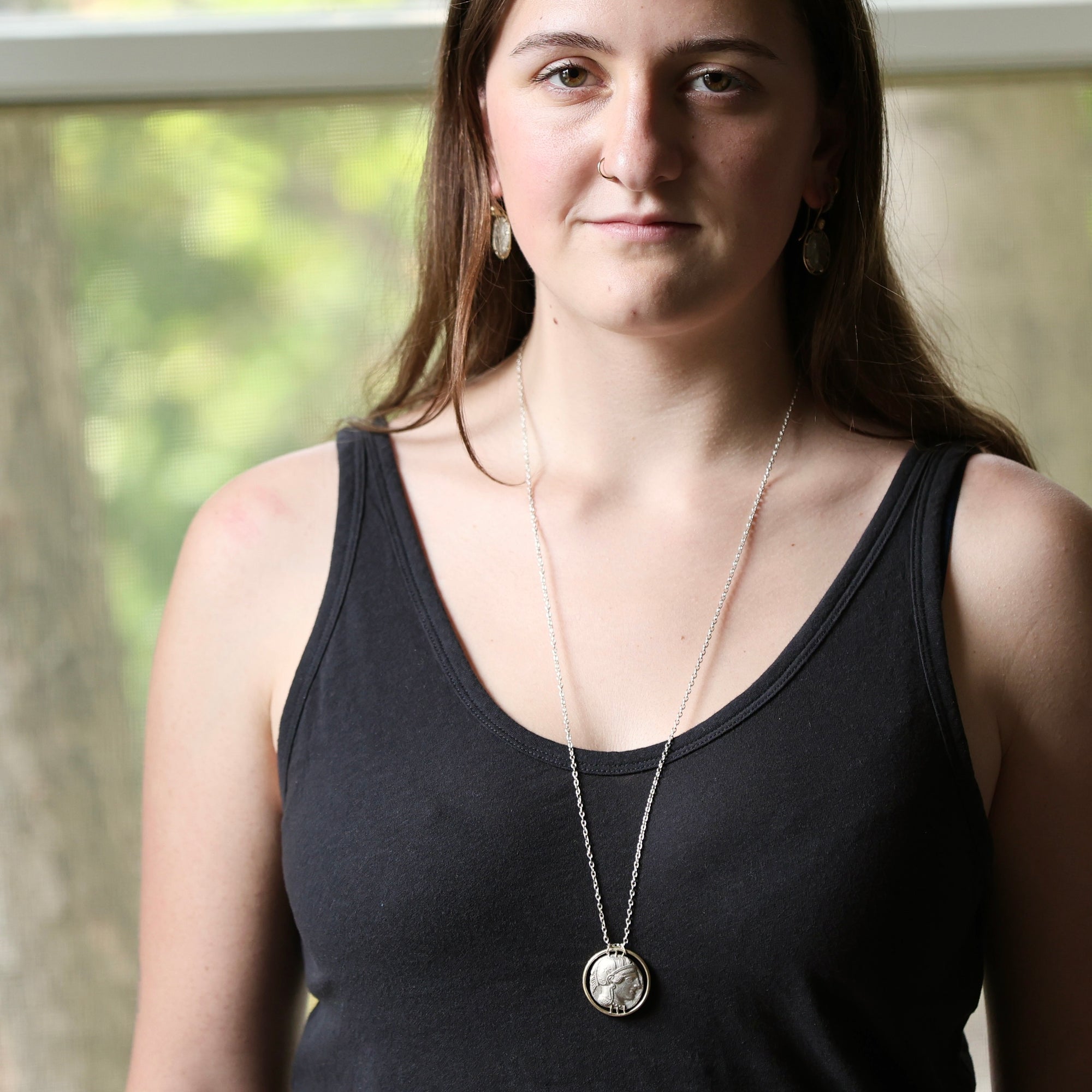 A woman wearing a silver coin pendant necklace and a black tank top, photographed from the chest up against a softly blurred window background.