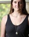 A woman wearing a silver coin pendant necklace and a black tank top, photographed from the chest up against a softly blurred window background.