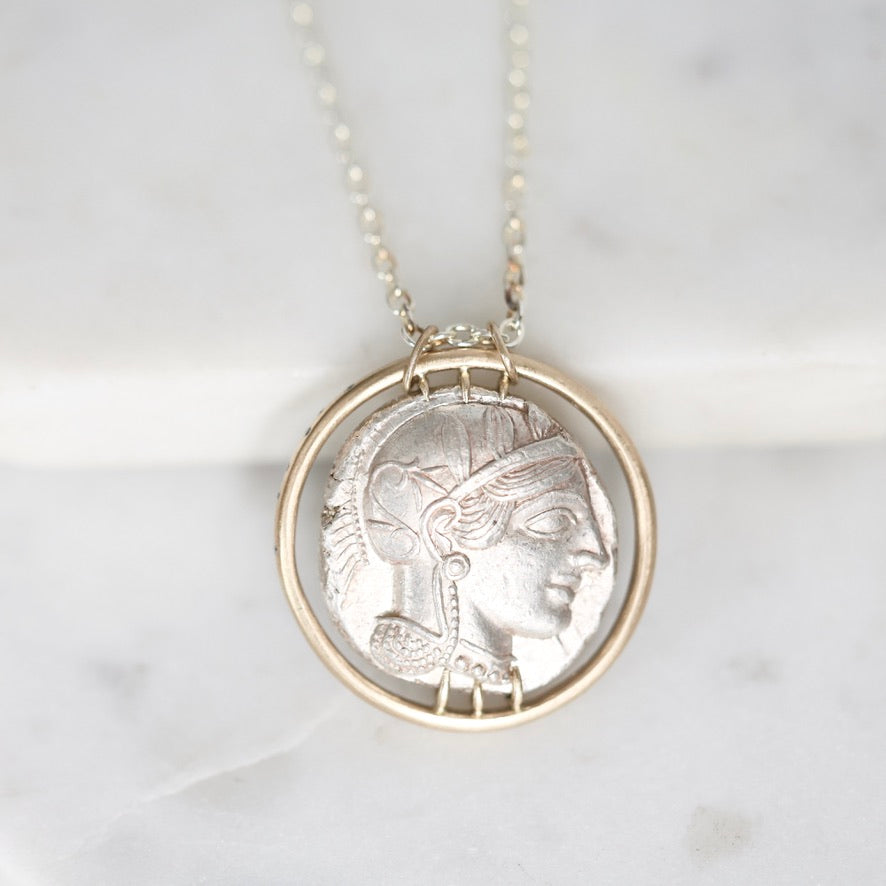 A silver pendant necklace featuring an ancient coin-style profile of a woman's face, set within a circular frame and hanging from a delicate silver chain on a light marble background.