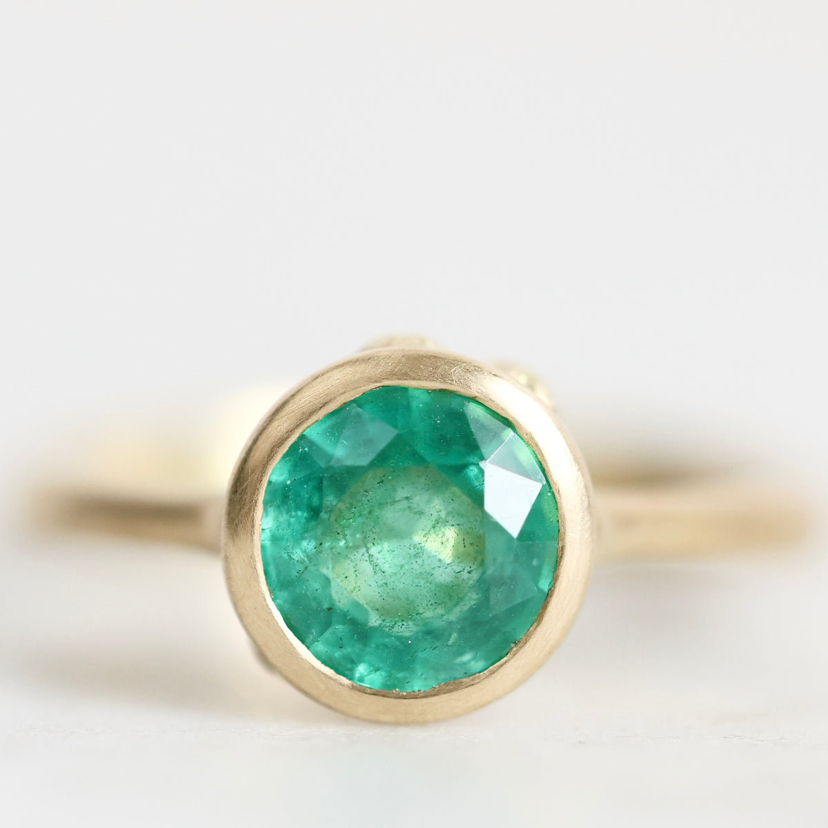 A gold ring featuring a round, faceted green emerald-cut gemstone in a prong setting, shown against a soft, light gray background.
