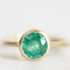 A gold ring featuring a round, faceted green emerald-cut gemstone in a prong setting, shown against a soft, light gray background.