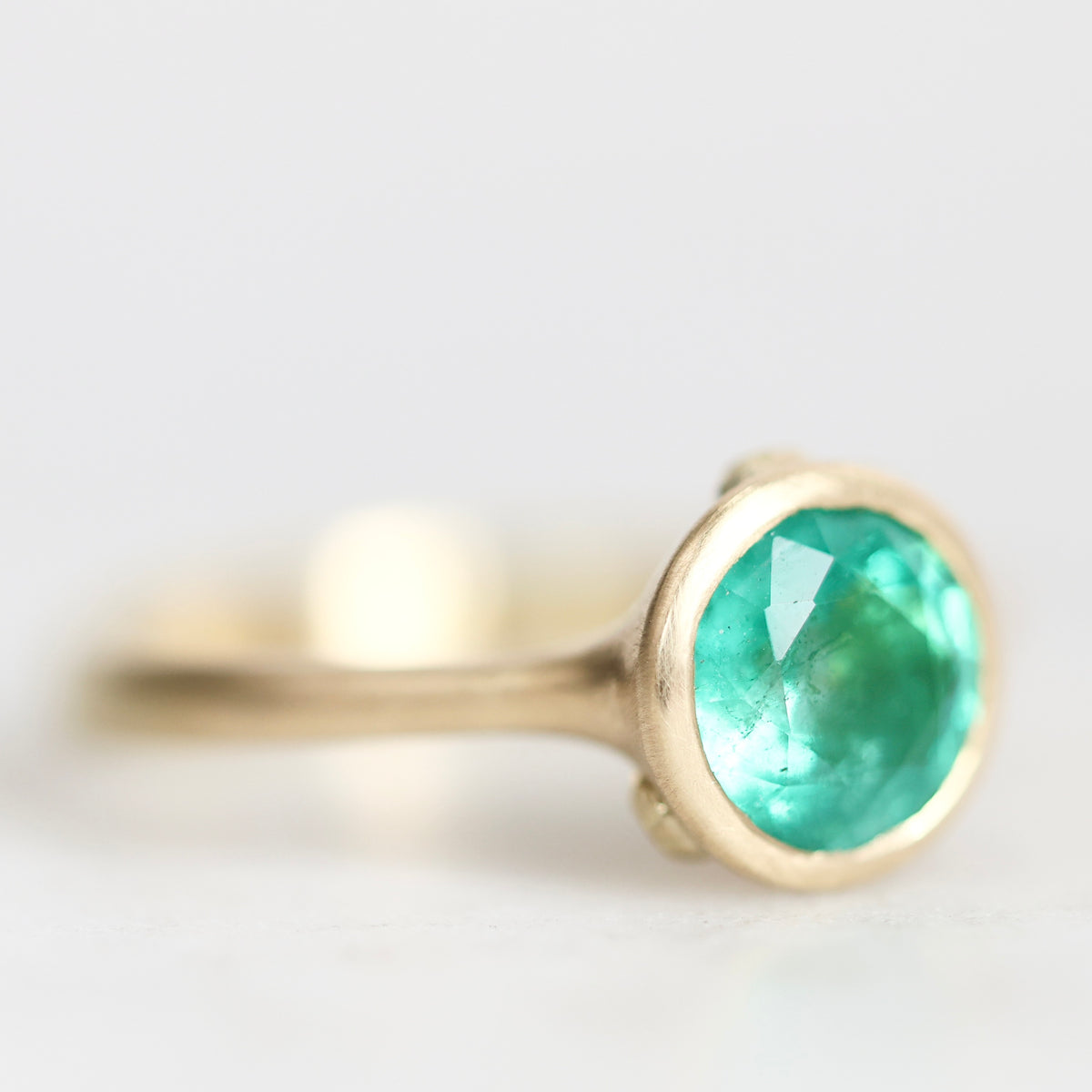 Gold bezel set ring with bright emerald green gemstone turned to the side against a white background.