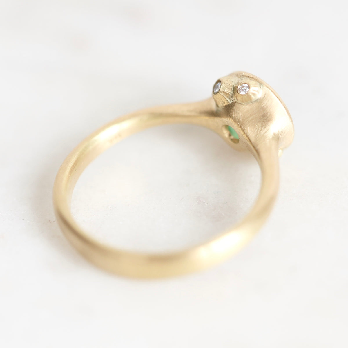 A gold ring with a sculpted, rounded head featuring two small clear gemstones and a tiny green accent, photographed on a light marble surface.