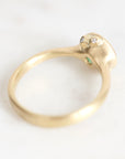 A gold ring with a sculpted, rounded head featuring two small clear gemstones and a tiny green accent, photographed on a light marble surface.