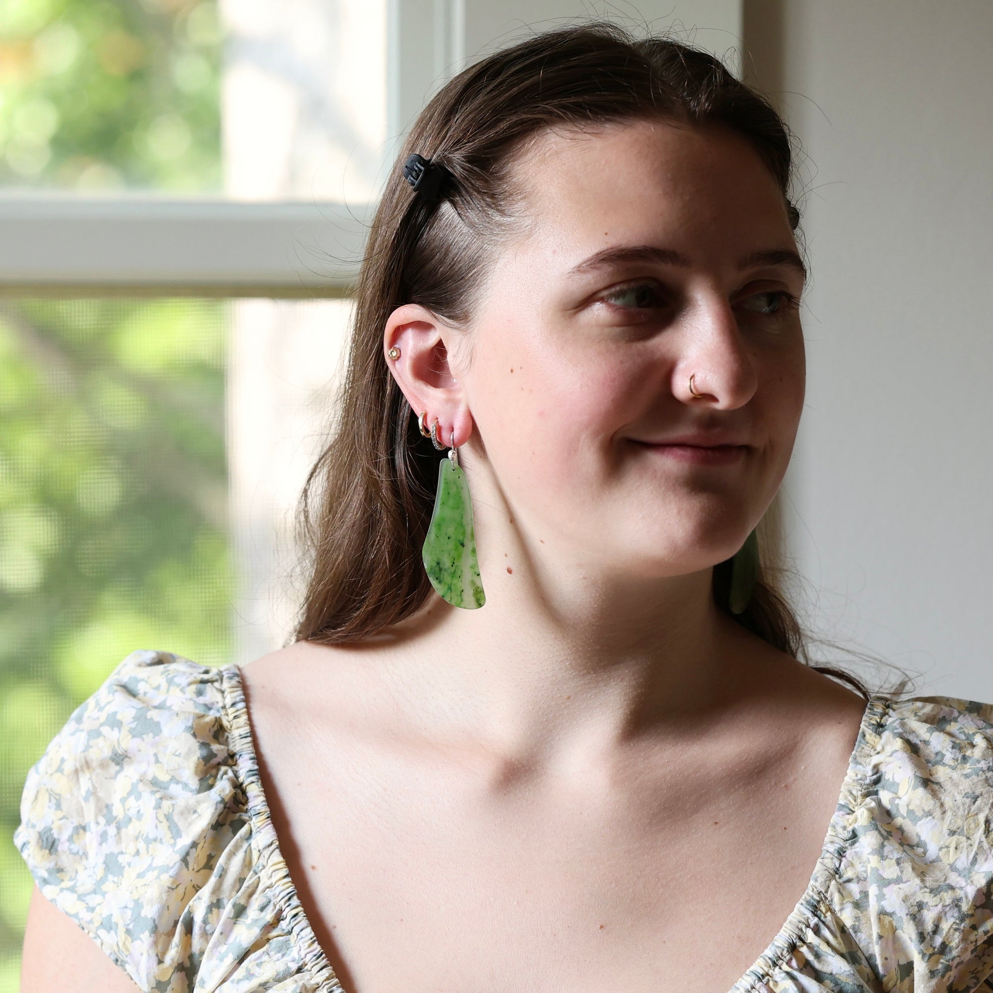A woman wearing a pair of green teardrop-shaped earrings with small black speckles, shown in a lifestyle portrait near a window.