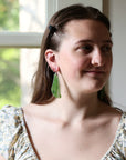 A woman wearing a pair of green teardrop-shaped earrings with small black speckles, shown in a lifestyle portrait near a window.