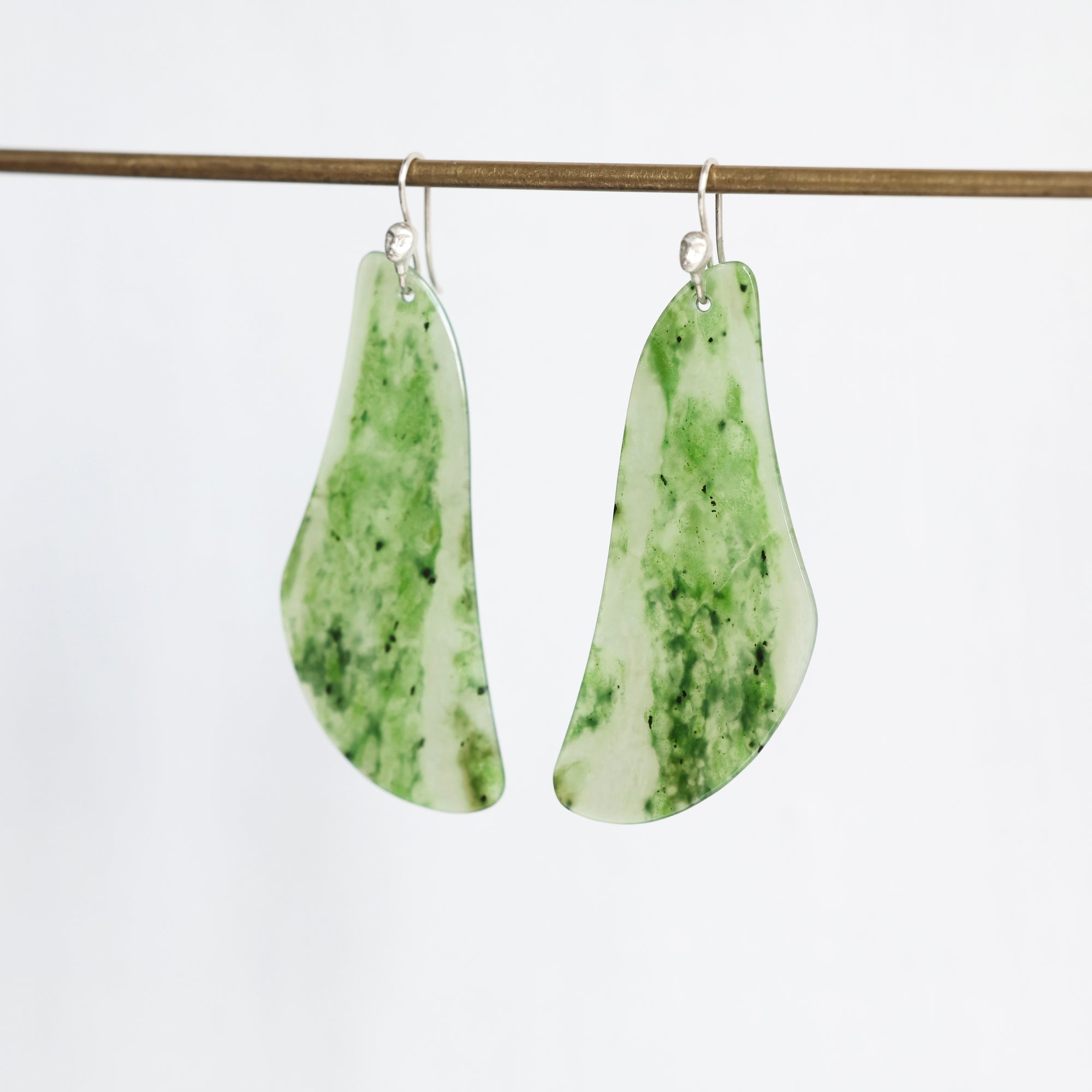 A pair of organic-shaped dangle earrings made from green and white marbled stone, hanging from silver fishhook wires on a thin wooden rod against a plain white background.