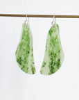 A pair of organic-shaped dangle earrings made from green and white marbled stone, hanging from silver fishhook wires on a thin wooden rod against a plain white background.