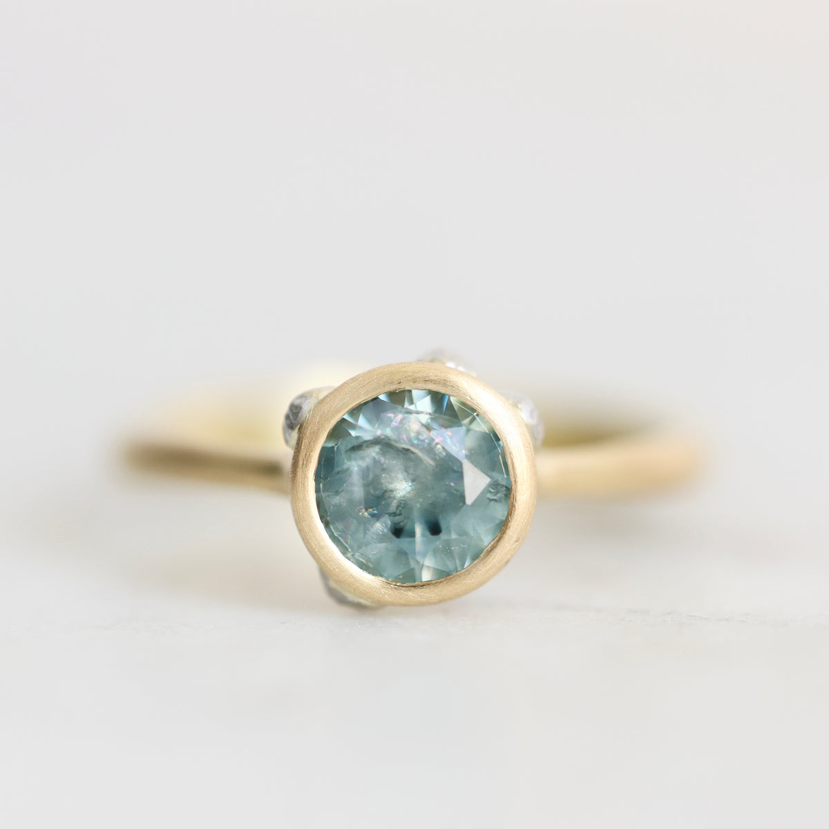 A close-up shot of a gold ring featuring a round-cut light blue gemstone in a bezel setting, with small clear accent stones on the shoulders, set against a soft, light gray background.