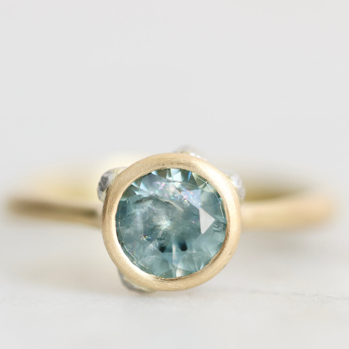 A close-up shot of a gold ring featuring a round-cut light blue gemstone in a bezel setting, with small clear accent stones on the shoulders, set against a soft, light gray background.