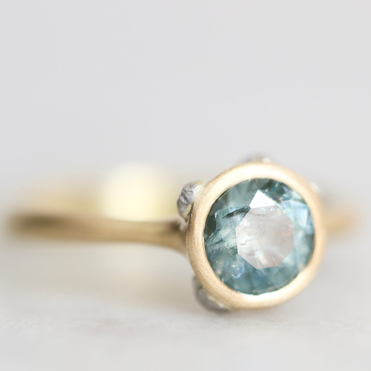 A close-up shot of a ring featuring a round faceted light blue gemstone in a gold bezel setting, with a silver-toned accent on the band, set against a soft, out-of-focus light gray background.