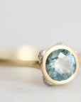 A close-up shot of a ring featuring a round faceted light blue gemstone in a gold bezel setting, with a silver-toned accent on the band, set against a soft, out-of-focus light gray background.